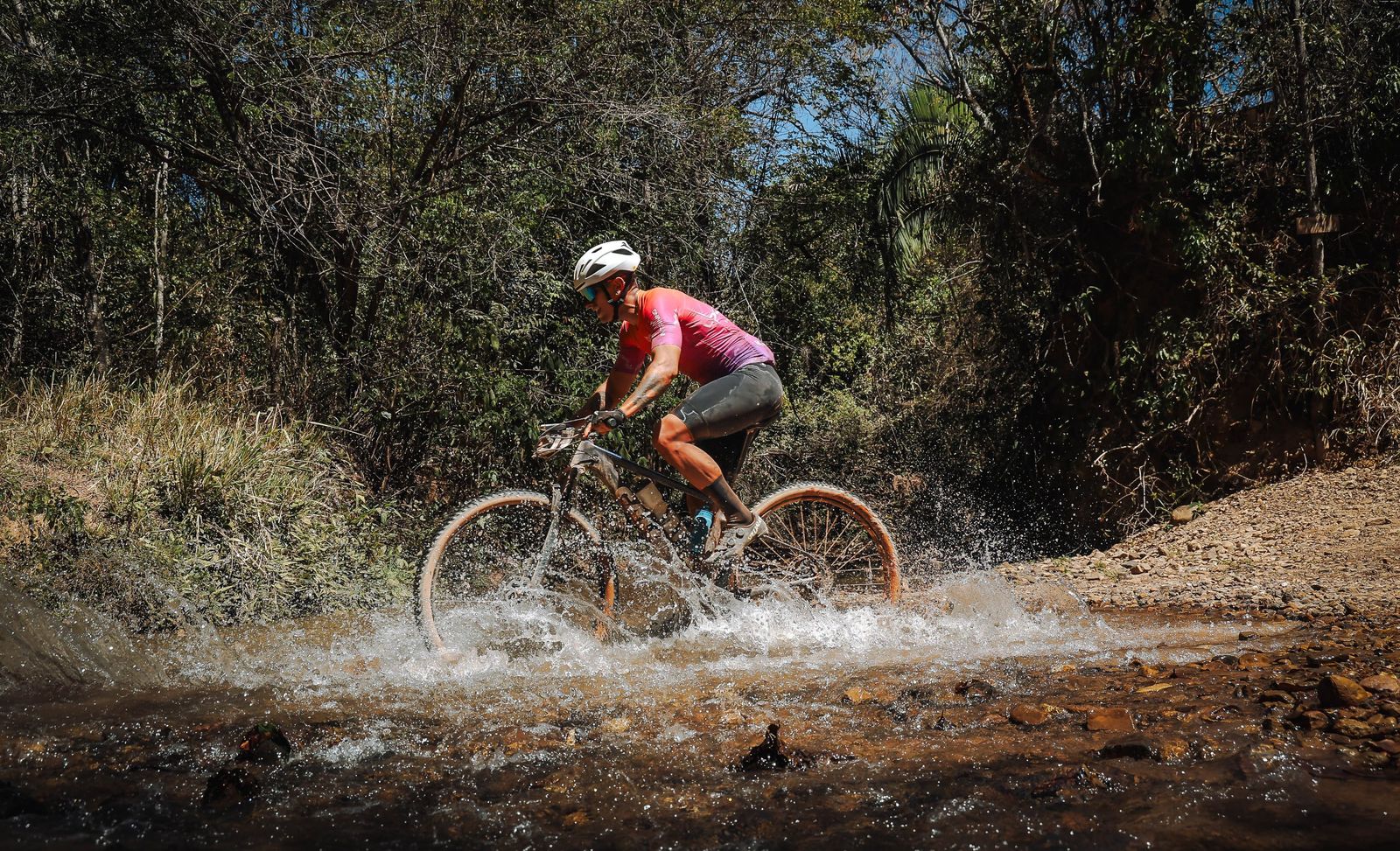 Bruno Paixão Dutra pedalando em treino de MTB, atravessando um curso d'água na trilha